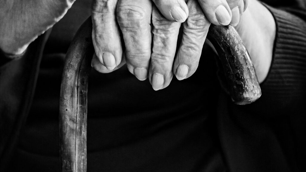A black and white close-up of an older person's hands resting on a walking cane